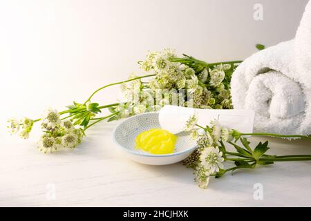 Kosmetische Feuchtigkeitscreme oder Weichmacher und ein Spatel für Pediküre und weiche Fußhaut in einer kleinen Schüssel und einige grüne Blumen auf einem hellen Holzhintergrund la Stockfoto