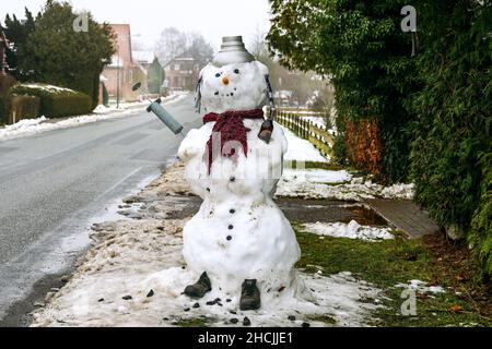 Carlow, Deutschland, 29. Dezember 2021: Auftauender Schneemann mit Impfspritze gegen Covid-19, Schal, Hut, Schuhe und eine Bierflasche auf einem ländlichen V aufgestellt Stockfoto