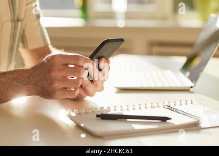 Horizontale Nahaufnahme eines nicht erkennbaren Mannes, der im modernen Büro am Schreibtisch sitzt und auf dem Smartphone im Internet surft Stockfoto