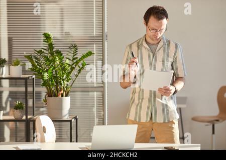 Horizontale, mittellange Aufnahme eines ernsthaften kaukasischen Mannes mit Brille, der im modernen Büro am Schreibtisch steht und durch die Papiere schaut Stockfoto