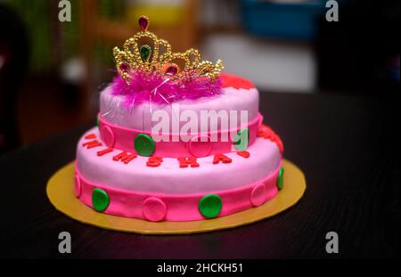 Zwei-Tier-Fondant-Kuchen-Design mit einer Tiara auf der Oberseite, mit Puderblumen verziert. Köstliche rosa-rote farblich thematisierte runde Torte auf gelbem Kuchenblech dagegen Stockfoto