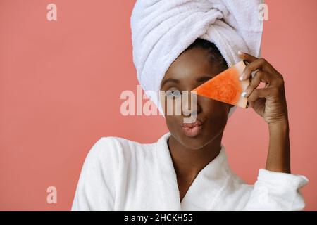 Junge schöne Frau lächelt mit einem umwickelten Handtuch um den Kopf, hält eine Scheibe Wassermelone und sendet Kuss auf rosa Hintergrund Stockfoto