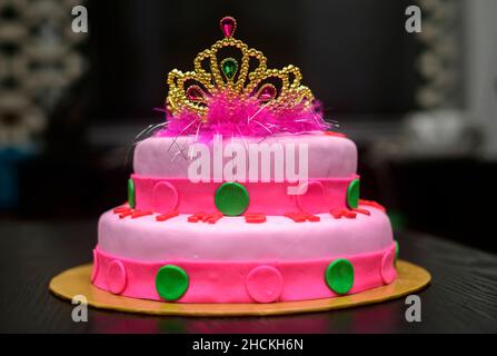 Zwei-Tier-Fondant-Kuchen-Design mit einer Tiara auf der Oberseite, mit Puderblumen verziert. Köstliche rosa-rote farblich thematisierte runde Torte auf gelbem Kuchenblech dagegen Stockfoto