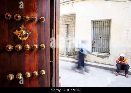 Kunstvoll verzierte Tür und Einheimische in einer engen Gasse in Stone Town, Sansibar, Tansania Stockfoto