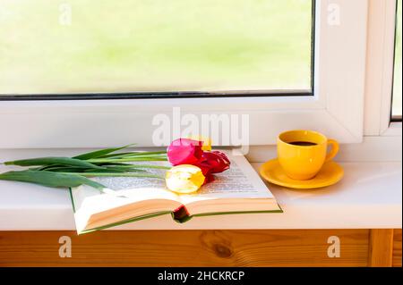 Geschnittene Tulpen liegen auf einem offenen Buch. Daneben auf der Fensterbank eine Tasse aufwachenden Kaffee Stockfoto