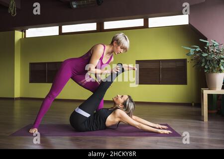 Der Personal Trainer hilft dem Schüler, eine Übung durchzuführen, um die Muskeln der Beine und des Rückens zu dehnen, er führt Training in Sportbekleidung auf einer Matte i durch Stockfoto