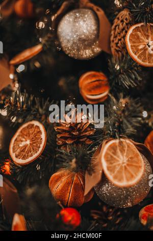 Getrocknete Mandarinen, Orangen, Zapfen, die an den Zweigen des Weihnachtsbaums hängen. Gemütliche Winterdetails und goldene Bokeh-Lichter. Dekorationen für den Winterurlaub Stockfoto