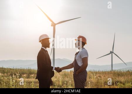 Afroamerikanischer Mann in weißem Helm und schwarzem Anzug, der mit einem indischen Ingenieur und Windturbinen im Hintergrund die Hände schüttelt. Zwei Kollegen, die sich über eine erfolgreiche Zusammenarbeit austauschen. Stockfoto