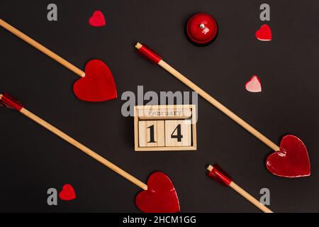 Draufsicht auf Holzkalender mit Schriftzug für den 14. februar in der Nähe von roten herzförmigen Lutschern auf Schwarz Stockfoto