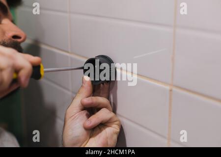 Mann Hände Befestigung Kunststoff-Komponente der Duschkopf Halter an gefliesten Wand im Waschraum mit Schraubendreher mit Teil des Gesichts im Hintergrund. Renovierung Stockfoto