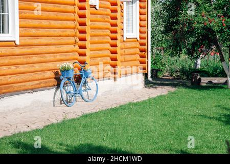 Sonnige Seite des hölzernen Landhauses mit dekorativem blauem Fahrrad mit Blumentopf auf Stamm, der links an der Hausfassade und Kirschbaum befestigt ist Stockfoto