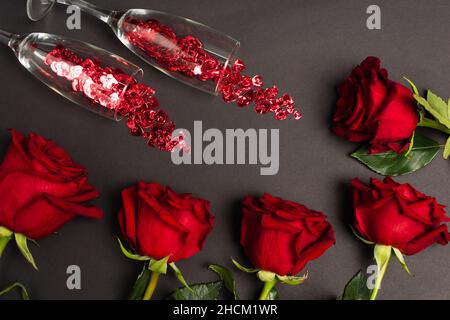 Draufsicht auf rote Rosen in der Nähe von Gläsern mit Konfetti auf Schwarz Stockfoto