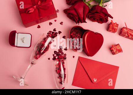 Ansicht von oben auf Champagner-Gläser mit glänzendem Konfetti neben Geschenkschachteln, Verlobungsring und roten Rosen auf rosa Stockfoto