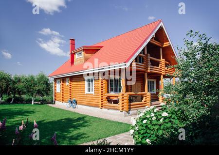 Tolles Holzhaus mit rotem Ziegeldach, Rasenfläche auf der linken Seite und Kirschbäume Obstgarten hinter dem Haus und vor Blumengarten im Vordergrund. Dorf Stockfoto