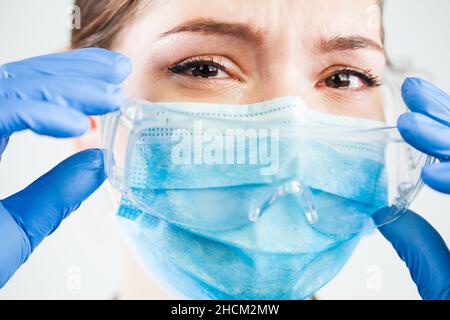Junge kaukasische Ärztin, die eine Schutzbrille einnahm, während sie Tränen der Trauer weinte, COVID-19 Coronavirus-Pandemie, die eine hohe Sterblichkeitsrate verursacht Stockfoto