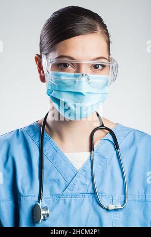 Porträt einer ernsthaften hübschen kaukasischen jungen Ärztin, trägt blaue Peelings Uniform, Schutzbrille, chirurgische Gesichtsmaske, Stethoskop Stockfoto