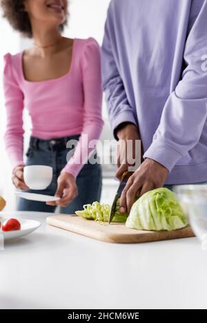 Beschnittene Ansicht des afroamerikanischen Mannes, der Kohl in der Nähe von verschwommener Freundin mit Tasse zu Hause schneidet Stockfoto