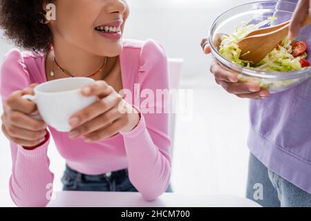 Beschnittene Ansicht einer afroamerikanischen Frau mit verschwommenem Becher, während der Freund zu Hause frischen Salat mischt Stockfoto