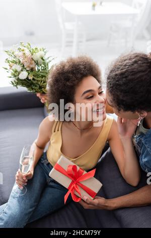 Blick aus der Ferne auf eine lächelnde afroamerikanische Frau, die Champagner in der Nähe des Freundes mit Blumen und Geschenkkarton zu Hause hält Stockfoto