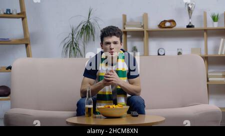 Besorgter junger Mann, der zu Hause ein Sportspiel im fernsehen in der Nähe von Bier und Chips ansieht Stockfoto