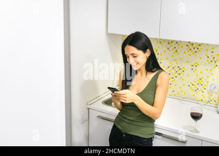 Junge kaukasische Frau hält ein Smartphone mit einem Smiley-Gesicht und Weinglas auf der Küchenoberseite Stockfoto
