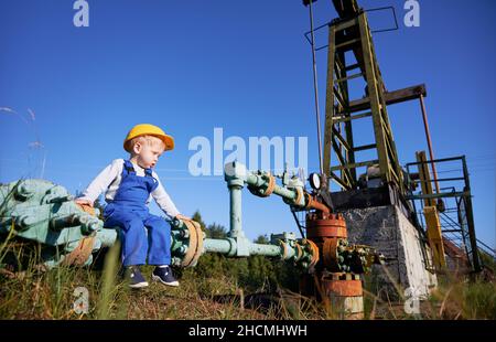 Kleiner Kindermechaniker, der auf Rohr der alten Ölpipeline in der Nähe des Erdölgutes sitzt und seine Struktur untersucht. Männliches Kind verbringt Zeit auf dem Ölfeld gegen den blauen Himmel. Stockfoto