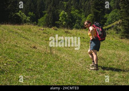 Attraktive Frau, die im Sommer an sonnigen Tagen mit Sonnenbrillen und Rucksack unterwegs ist und auf die Kamera blickt Stockfoto