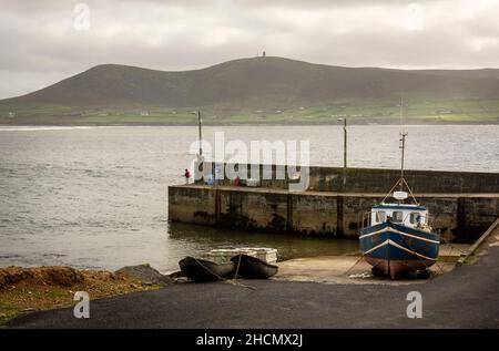 Der Pier am Rinroe Point, in der Nähe von Carrowteige, Irland. Ein Angler steht mit seiner Angelrute am Ende des Piers. Zwei Boote sind an Land. Stockfoto