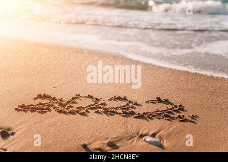 2022 Figuren am Sandstrand Frohes Neues Jahr 2022, Zahl Hand in den goldenen Sand auf schönen Sonnenuntergang goldenes Licht geschrieben. Die Meereswelle wäscht die ab Stockfoto