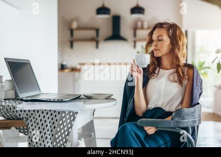 Frau sitzt vor einem Laptop am Küchentisch und schaut auf den Bildschirm. Arbeiten von zu Hause aus. Stockfoto
