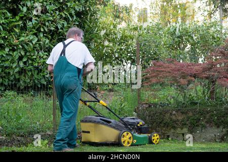 Mann, der im Frühjahr den Rasenmäher durch einen kleinen Hinterhof schiebt. Rückansicht des Menschen mit dem Gesamtbild. Stockfoto