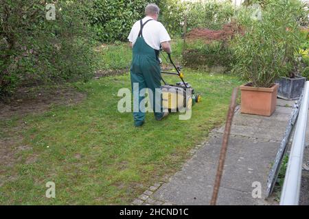 Mann, der im Frühjahr den Rasenmäher durch einen kleinen Hinterhof schiebt. Rückansicht des Menschen mit dem Gesamtbild. Stockfoto