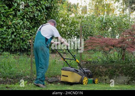 Mann, der im Frühjahr den Rasenmäher durch einen kleinen Hinterhof schiebt. Rückansicht des Menschen mit dem Gesamtbild. Stockfoto