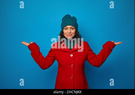 Konzeptuelles Porträt einer schönen jungen, lockigen Brünette in rotem Mantel und grünem Hut posiert vor blauem Hintergrund und blickt auf die Kamera, die hält Stockfoto