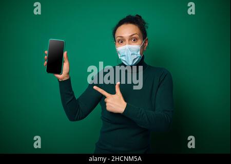 Überraschte junge tausendjährige Frau in schützender medizinischer Maske schaut auf die Kamera und zeigt ihren Finger auf ihr Smartphone mit leerem leeren Bildschirm für Kopie s Stockfoto