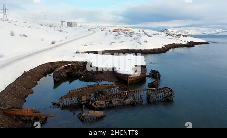 Luftaufnahme der schneebedeckten Küste mit zerstörten Booten nach dem Schiffswrack. Alte versunkene Fischerboote am Ufer der Barentssee. Stockfoto