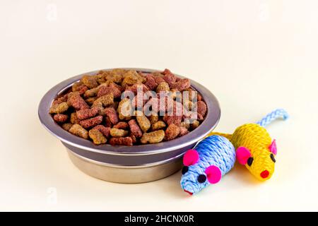 Gesundes trockenes Katzenfutter. Braune knusprige Bio-Kitty-Kibble-Stücke in einer Schüssel für Tierfutter und Maus-Spielzeug auf hellem Hintergrund. Stockfoto