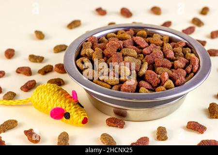 Gesundes trockenes Katzenfutter. Braune knusprige Bio-Kitty-Kibble-Stücke in einer Schüssel für Tierfutter und Maus-Spielzeug auf hellem Hintergrund. Stockfoto