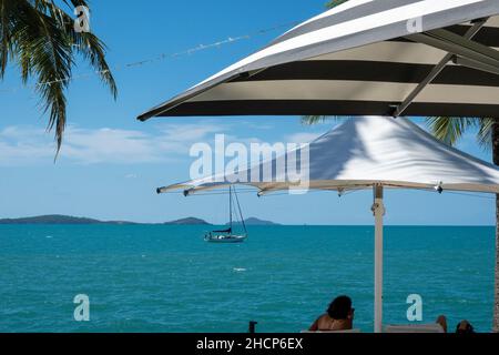 Touristen entspannen sich unter Sonnenschirmen und schauen auf eine Yacht, die vorbei segelt Stockfoto