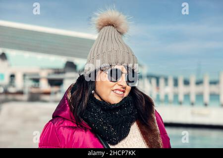 Porträt einer lächelnden Frau mit Sonnenbrille und Wollmütze an einem sonnigen Herbsttag, der durch die Stadt geht Stockfoto