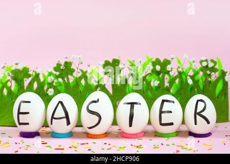 Weiße Eier mit schwarzen Buchstaben OSTERN und Gras auf einem rosa Hintergrund. Frohe Ostern Karte. Flach liegend, Draufsicht, Kopierbereich. Stockfoto