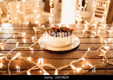 Weißer Hochzeitstorte mit Früchten auf einem Holztisch, der mit Blumengirlanden geschmückt ist Stockfoto