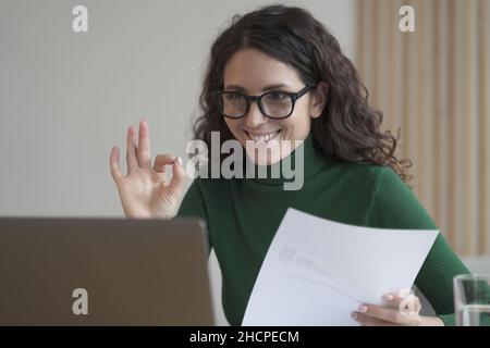 Lächelnde italienische Mitarbeiterin, die den Finanzbericht hält und während eines Videoanrufs eine gute Geste zeigt Stockfoto