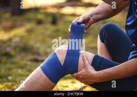 Erste Hilfe nach einem Wanderunfall. Verletzte Wanderin, die ihr elastische Binde auf das Knie legte. Sehnenprobleme oder Verstauchungen des Knies während des Trekkings in der Natur Stockfoto