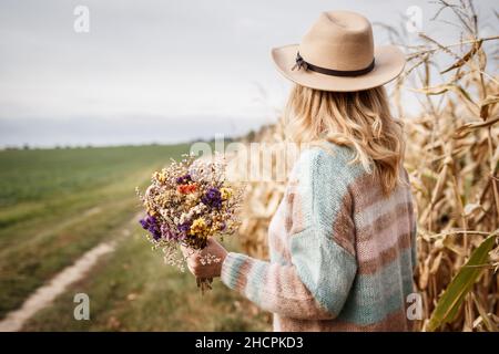 Frau mit gestricktem Pullover und Hut, die einen Strauß getrockneter Blumen im Freien hält. Kräuter und blütengetrocknete Pflanze in weiblichen Händen. Ländliche Landschaft Stockfoto