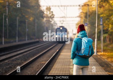 Frau mit Rucksack schaut auf den ankommenden Zug an einem Bahnhof. Reise- und Transportkonzept Stockfoto