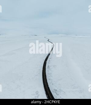 Luftdrohne Blick bergauf Straßenlandschaft im Winter auf Island. Asphaltstraße mit seitlichem Schnee im Winter mit blauem Himmel. Stockfoto