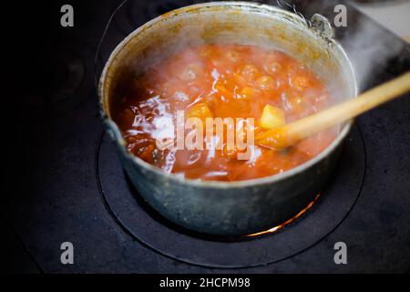 Gusseiserner Kessel kocht einen Gulasch-Eintopf über einem Holzofen aus roten Ziegeln im Hinterhof eines ländlichen Hauses in Rumänien. Stockfoto