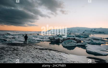 Junge Touristen stehen vor dem berühmten See Joekulsarlon Gletscherlagune und Diamantenstrand mit seinen Eisbergen und Eisschollen in Island während des Sonnenuntergangs Stockfoto