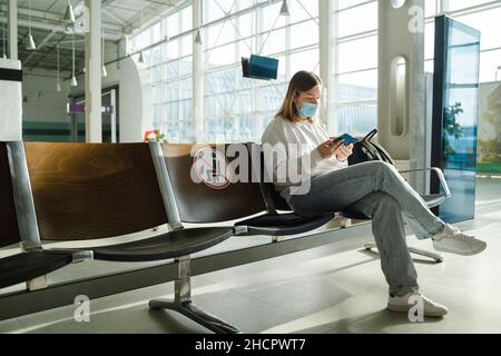 Weibliche Touristen in Maske sitzen in der Lounge des Flughafenterminals auf leeren, gekennzeichneten Stühlen unter den neuen normalen und aufrechten sozialen Fernabstandsbestimmungen. Sitzende Frau Stockfoto